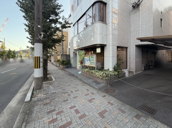 歩道から見上げたビルの角。曲面ガラスの窓が上階へ重なり、1階には郵便局の掲示板と花壇、左手に車道と電柱が並ぶ