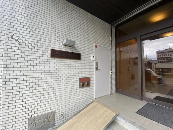 入口脇の白いタイル壁に掛かる「三善ビル」の銘板と白い扉、足元の定礎石。右手にガラスの自動ドアが見える