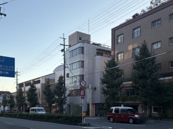 交差点の向かいから見た左京区の白川森ビル。白いタイル張りの細長い建物で、角の丸い窓と屋上の塔屋が茶色の建物の隣に見える