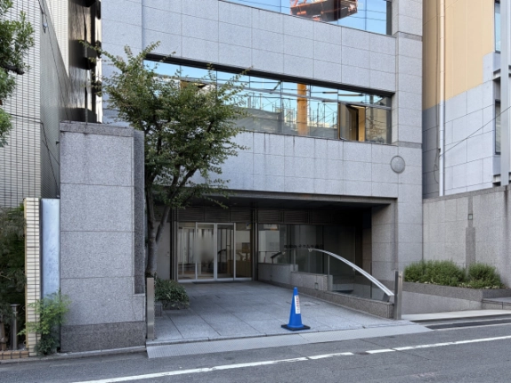 石敷きのアプローチの奥にある自動ドアのエントランス。手前左に植栽の木、右に曲線のステンレス手すり