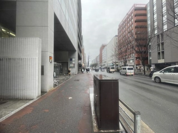 建物を左手に見た前面道路。奥へ続く歩道と街路樹、通り向かいのビル群