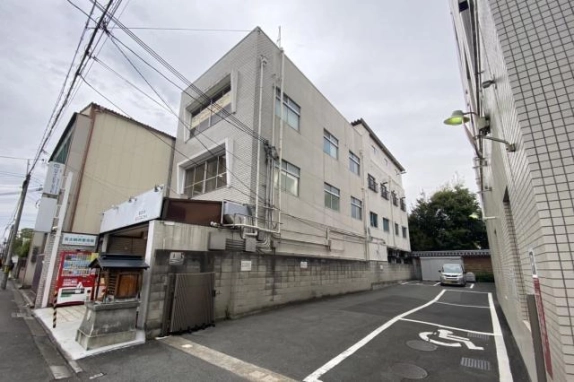 隣の駐車場側から見上げた3階建ての側面外観。白い外壁に各階の窓が並び、1階の脇にブロック塀と門扉がある