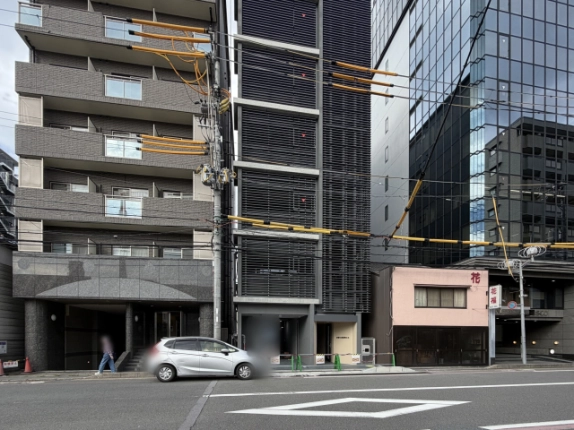 通りの向かいから見た下京区の街並み。左のタイル張りマンションと、右の低層建物やガラス張りのビルに挟まれた細長いルーバー外観のビル