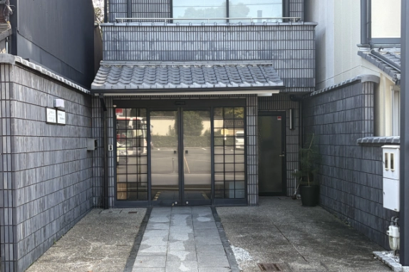 1階入口の近景。瓦の庇の下に格子入りのガラス扉、右側に別の出入口ドアがあり、石畳の通路の両側に前面空地が広がる