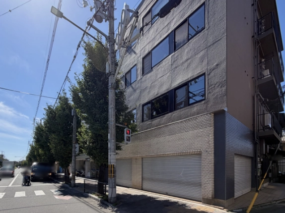 電柱と歩行者用信号越しに見た建物正面。上階は黒いサッシの横長窓、1階は白いレンガ調タイル壁にシャッターが並ぶ