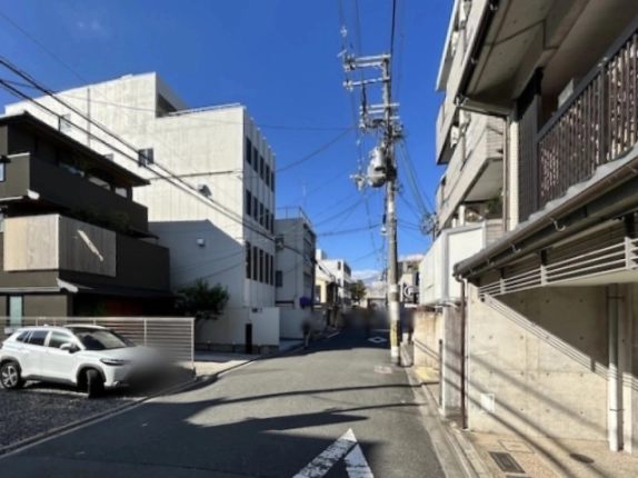 前面道路を反対方向から見た街並み。左手の黒い外壁の住宅の奥に、縦長の窓が並ぶ白いテナントビルが建つ