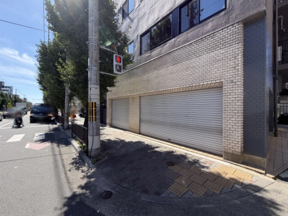 角の歩道から見た1階。幅の異なる2枚のシャッターと、点字ブロックのあるレンガ敷きの歩道