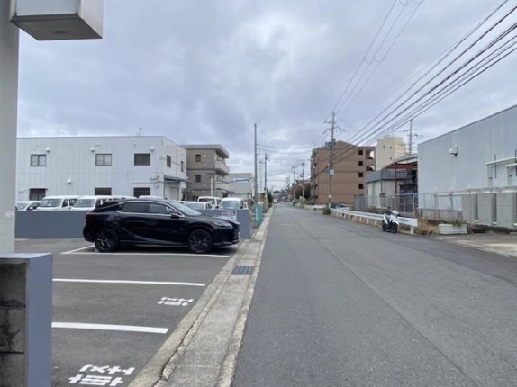 前面道路の様子。左に車が停まる駐車場、右に白い倉庫風の建物と電柱が並び、道がまっすぐ奥へ続く