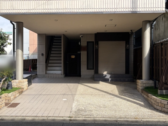 1階ピロティ奥の近景。左に上階へ続く階段と黒い格子扉、右に両脇に照明が付いたシャッターの区画