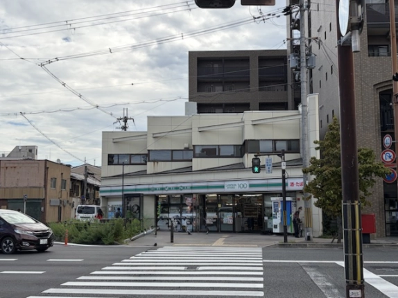 交差点の横断歩道越しに見た建物と周辺。白い2階建ての奥に高層の建物が立つ
