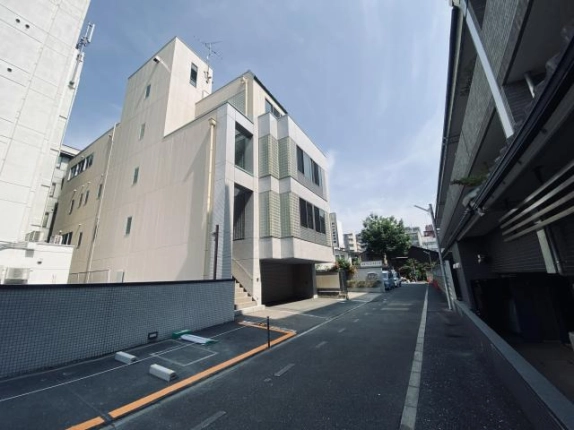 道路側から見たビルの側面。屋上にアンテナ付きの塔屋が立ち上がり、手前にタイル塀沿いの駐車スペース、右手に隣の建物が並ぶ