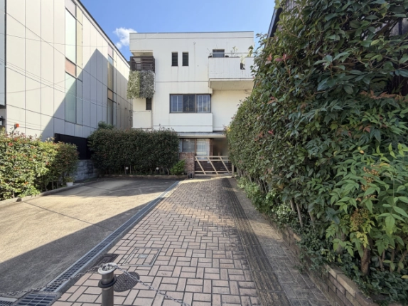 通路手前から正面に建物を望むアプローチ。右側に背の高い生垣、左側にコンクリート舗装の敷地と排水溝が続く