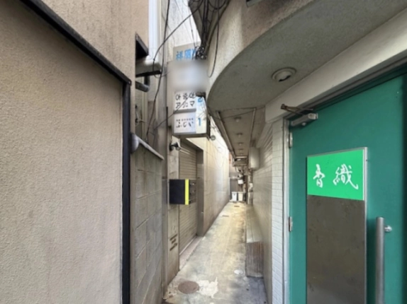 祥朋ビルの袖看板が縦に並ぶ路地。右手前に緑色の扉、左奥にシャッターと集合ポストがある