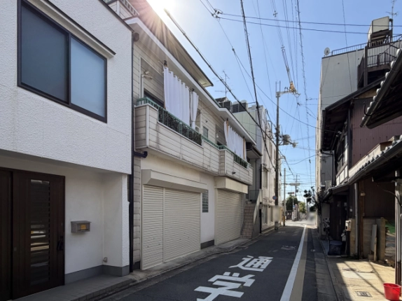 上京区の細い通りに建つ様子が分かる街並み。白い住宅の隣に建ち、路面に「通学路」の表示、向かいに町家が並ぶ