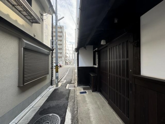 町家の入口前から通りへ向かう路地。右手に木格子の戸、左手に向かいの建物