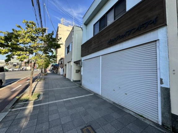 建物に近づいて見た1階の2連シャッターと木目の看板帯。左奥へタイル敷きの歩道と街路樹が続く