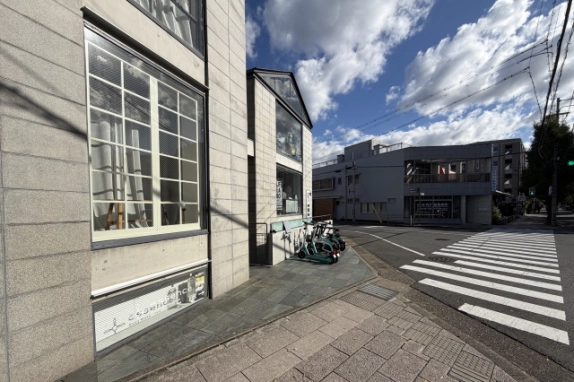 建物の角にある白い格子の大きな窓と石張りの歩道。壁際に電動キックボードが並び、右手に横断歩道がある