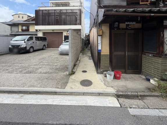 下京区の通りから見た路地の入口。左に車が停まる駐車場、右に格子戸の町家があり、その間の細い通路が奥へ延びる