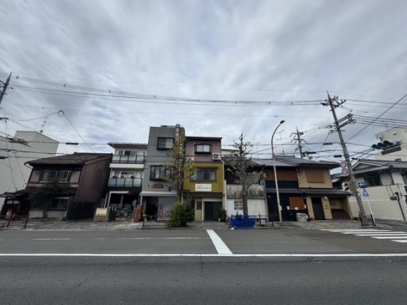 東大路通の向かい側から見た東山区本瓦町の街並み。中央の黄色い外壁の3階建てが本物件で、右手に横断歩道と信号がある