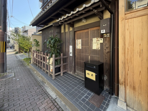 玄関まわりの様子。木製の格子戸の前はタイル張りで、右に黒い宅配ボックス、左に木柵と鉢植えが置かれている