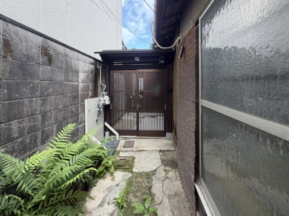 路地の奥にある小屋根付きの格子引き戸。手前にはシダが茂るひび割れた通路があり、左にブロック塀、右に型板ガラスの窓が並ぶ