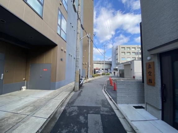京都駅徒歩5分の新築3階建一棟貸店舗の周辺道路。左に大型建物の出入口と電柱、右奥にカラーコーンの並ぶ舗装スペース、正面奥に高架が見える