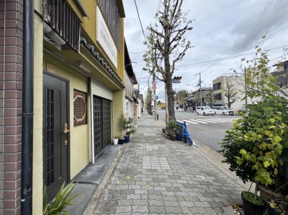 少し離れて見た1階入口と前面の歩道。石畳風の舗装と街路樹が続き、車道の先に横断歩道がある