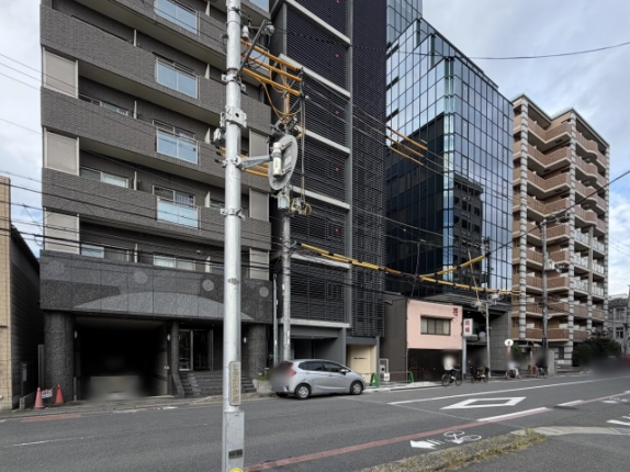 手前の電柱越しに左斜めから見た建物と前面道路。隣のマンションや右奥のガラス張りの高層ビルとの位置関係がわかる