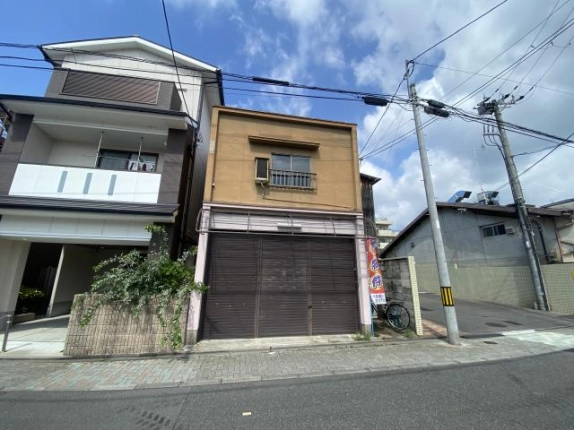 通りの向かい側から見た2階建ての建物。左に3階建ての住宅、右に電柱と舗装された敷地があり、手前にブロック舗装の歩道と車道が通る