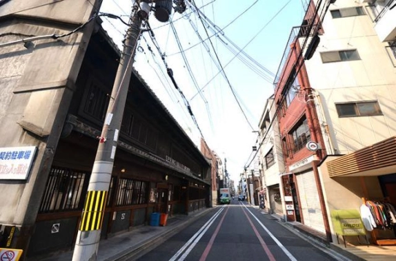 建物前の通りの眺め。左側に木格子の町家が続き、右手に赤い外壁の本物件と白いビルが並ぶ