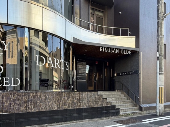 「KIKUSAN BLDG」の看板があるエントランスの近景。ガラス扉の脇にテナント案内板があり、手すり付きのタイル階段で入口へ上がる