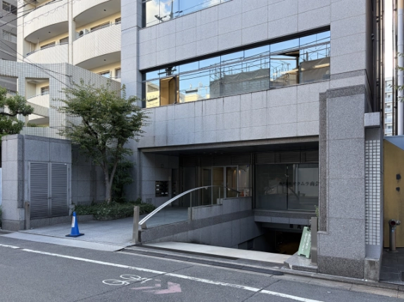 斜め前から見た石張りのビル低層部。左に植栽と白い集合住宅、中央から右に曲線の手すりが付いた地下への車路