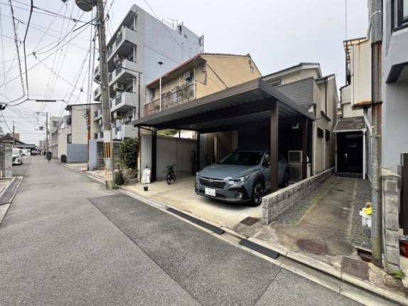 道路側から見た敷地まわり。中央にカーポート、右端に奥の玄関へ続く細い通路があり、背後に中層マンションが建つ