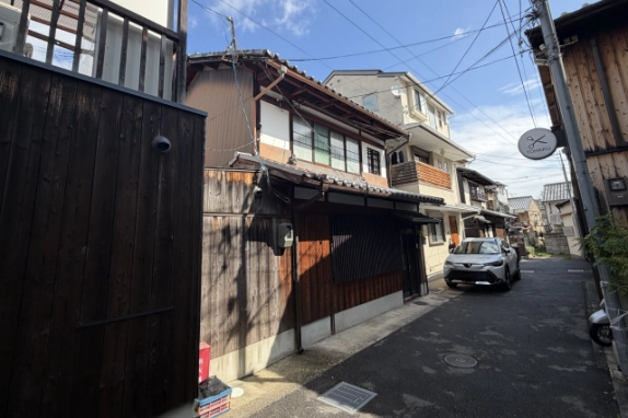 路地の手前から見た京町家と周辺の住宅。黒い板塀の奥に町家の瓦屋根と格子窓が見え、路地の先に白い車が停まっている