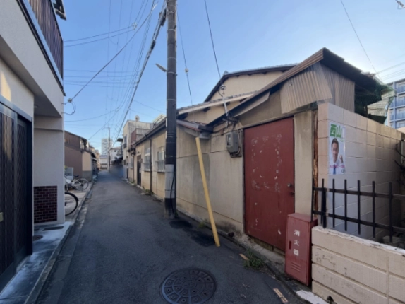 周辺の路地。右手に赤い鉄扉とトタン屋根の建物、黄色い支線カバー付きの電柱、手前にマンホール