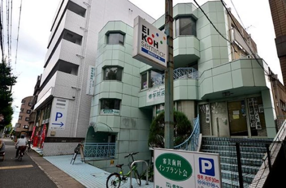 左京区の通り沿いから斜めに見た外観。手前に電柱の看板と自転車、右に水色の手すりが付いたタイル張りの外階段、左に歩道と車道が続く