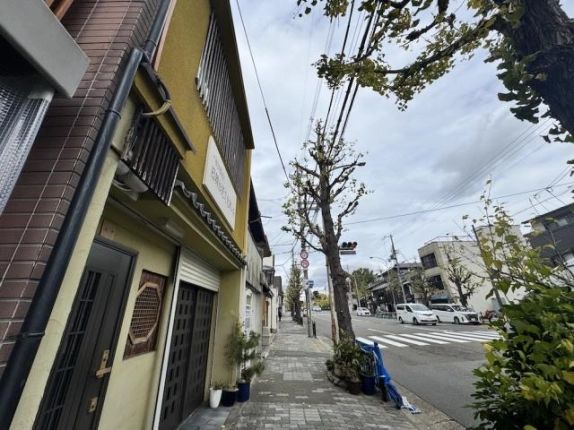 見上げるように撮影した外観。黄色い外壁の2階に縦格子の窓と看板があり、右手に東大路通の街路樹と電線が広がる