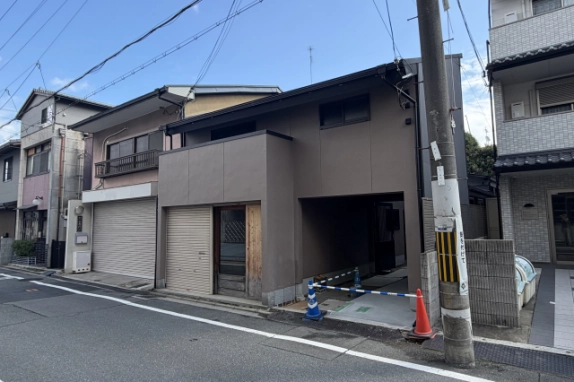 斜め右から見た建物外観。2階に窓が並び、1階右側の通路前にカラーコーンとバーが置かれ、手前に電柱が立つ
