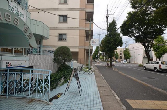 修学院駅徒歩7分の建物入口前から見た前面道路。左に水色の装飾手すりと立て看板、右に並木の続く車道と先の横断歩道が見える