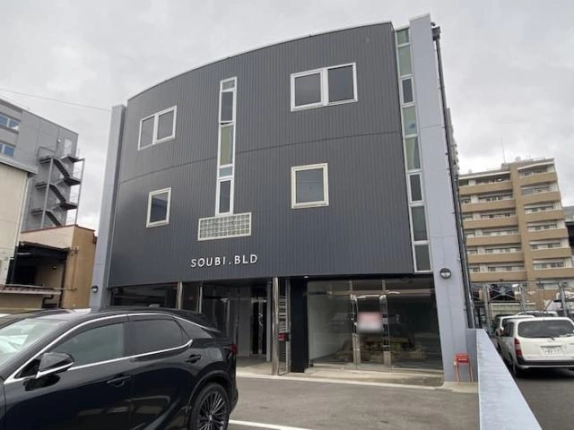 右斜め前から見た鉄骨造3階建のビル外観。白い窓枠の窓とガラスブロックが並び、1階に入口とガラス張りの区画、右隣にマンションが見える