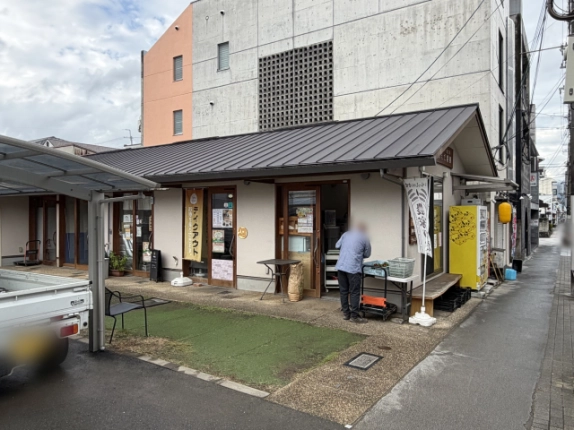 北区の道路沿いから見た平屋の店舗棟。金属屋根の下に店舗の入口が並び、手前に人工芝のスペース、右に黄色い自動販売機