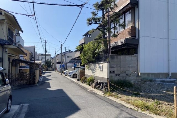 建物の脇から見た前面道路。住宅が並ぶ細い道で、右手に松の木とレンガ調・白い外壁の建物