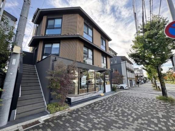 建物左手に2階へ直接上がれる外階段があり、隣にガラス張りの1階店舗、右手に川端通の歩道と街路樹が続く外観
