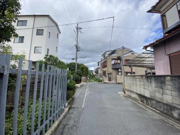 右京区太秦松本町の住宅街の道路。左側のグレーの柵越しに白い建物が見え、右側には住宅とブロック塀が続く