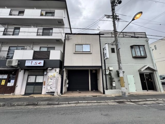 道路の向かいから見た正面外観。西京極駅徒歩12分の2階建てで、5階建てマンションとタイル張りの建物に挟まれ、1階に黒いシャッターが並ぶ