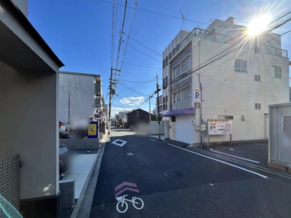 物件前の道路を見通した様子。路面に自転車マークがあり、右手に紫色の庇の隣接ビルとコインパーキングが見える