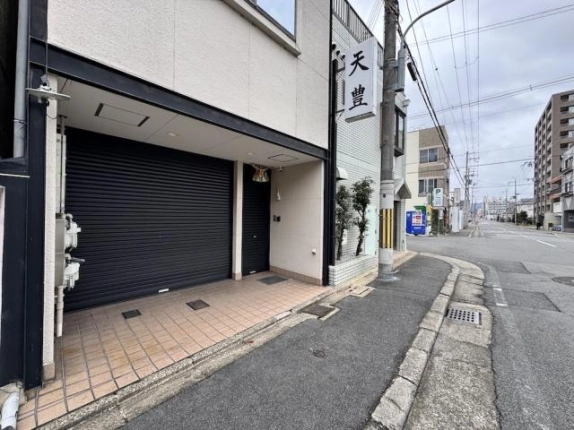 右京区西京極南庄境町の1階テナントを右斜め前から見た外観。黒いシャッターとタイル張りの入口前があり、右手に歩道と道路が続く