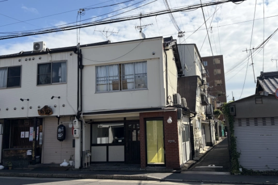 やや右手前から見たカフェ居抜きの1階店舗。木製扉と赤レンガ部分の右側に、奥へ続く細い路地がある