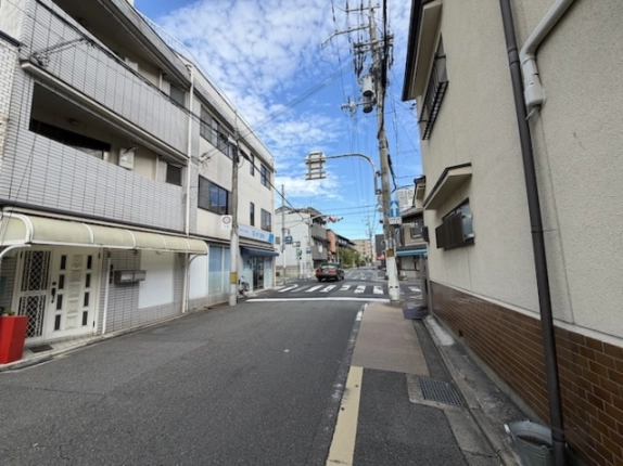 ビル前の道路を交差点の方向に見た様子。左手にタイル張りの隣の建物とビルの1階、正面奥に横断歩道と信号機が見える