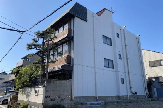 鉄骨造3階建ての側面。白い外壁に小窓と縦樋が並び、左手にレンガ調の正面と松の木が見える